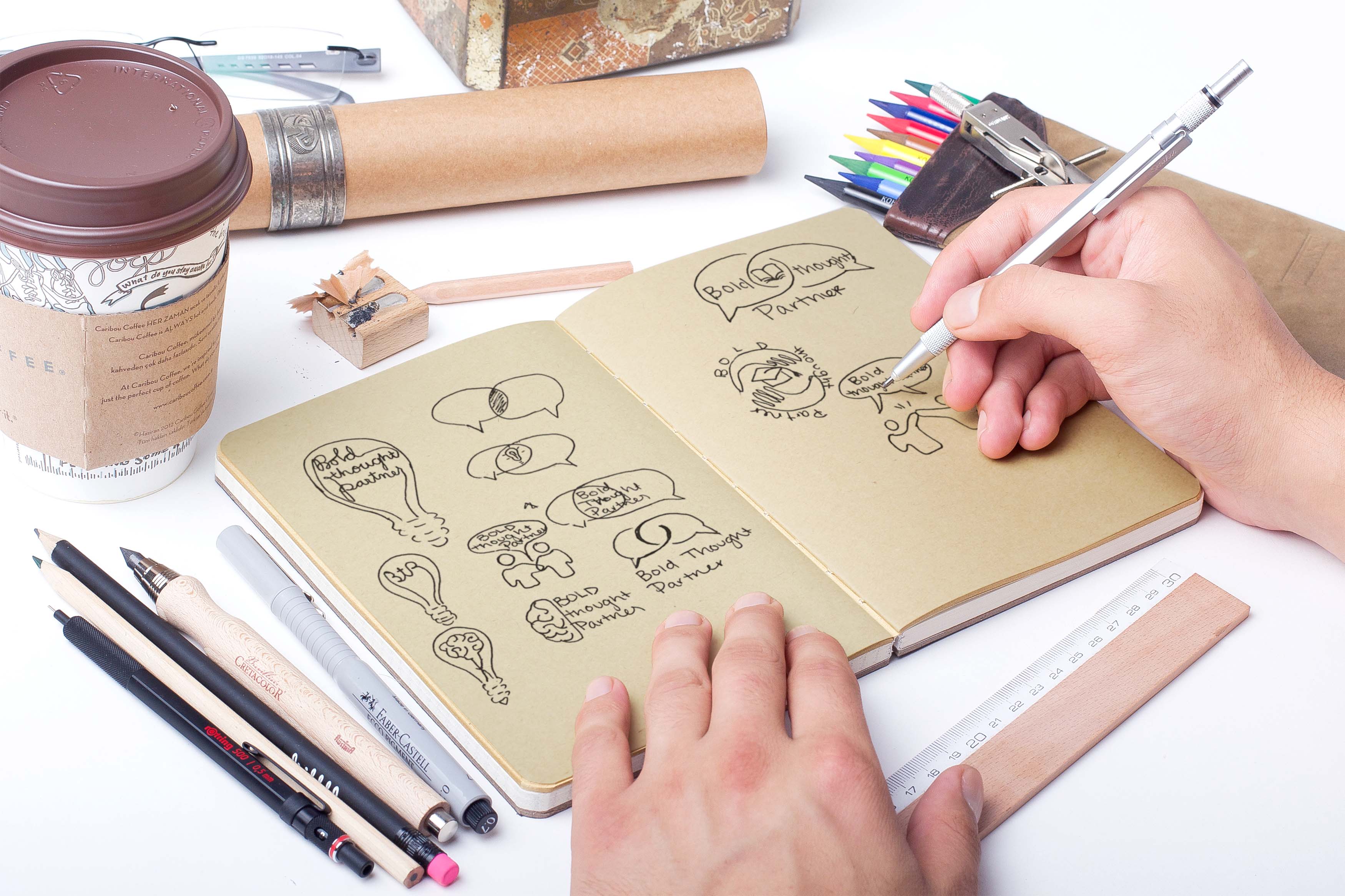 Designer sketching on a sketchpad with various supplies sitting on desk near the sketchbook.