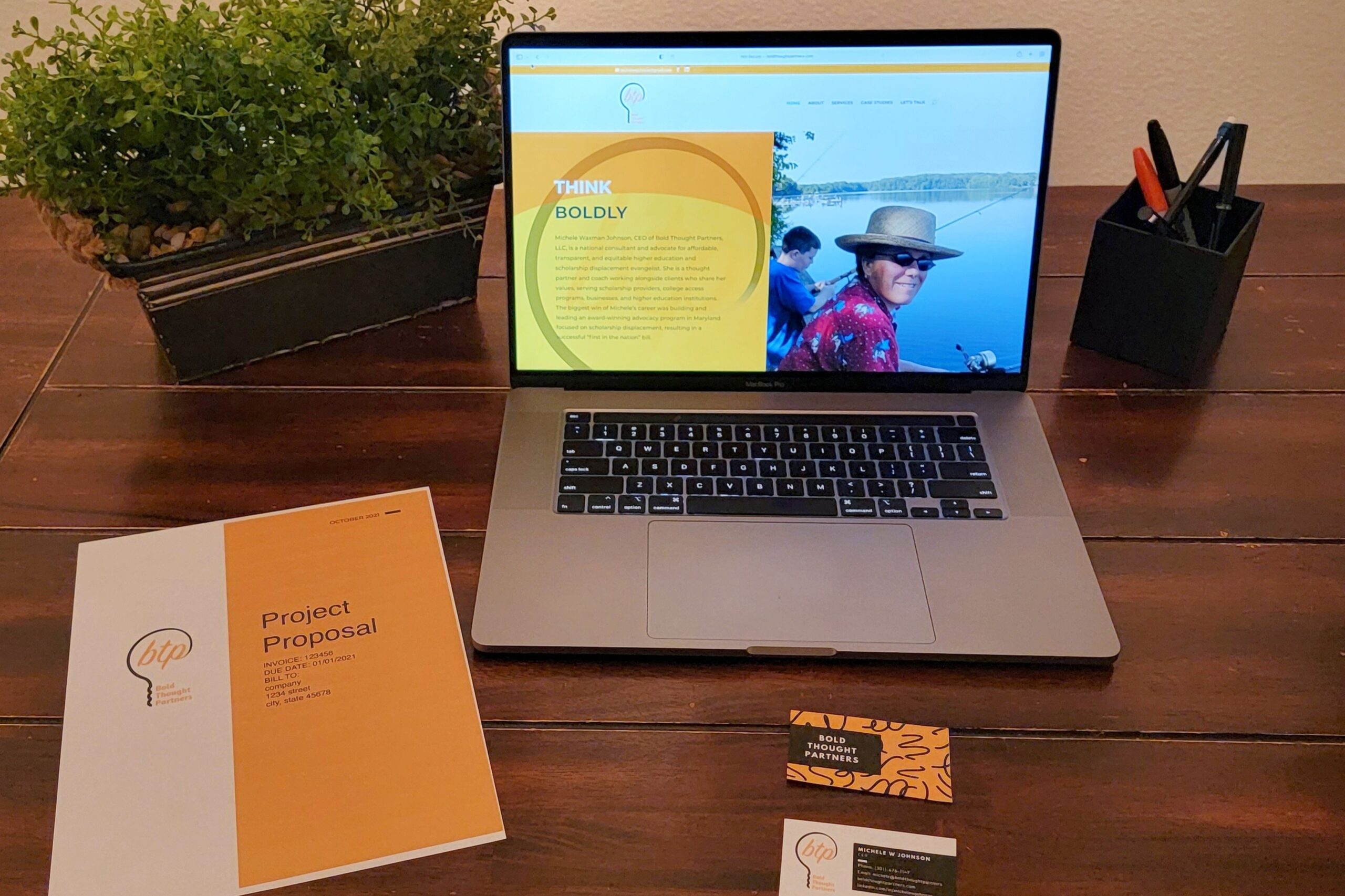 A desk with a plant, pen cup, open laptop, proposal and business cards layed out on top.